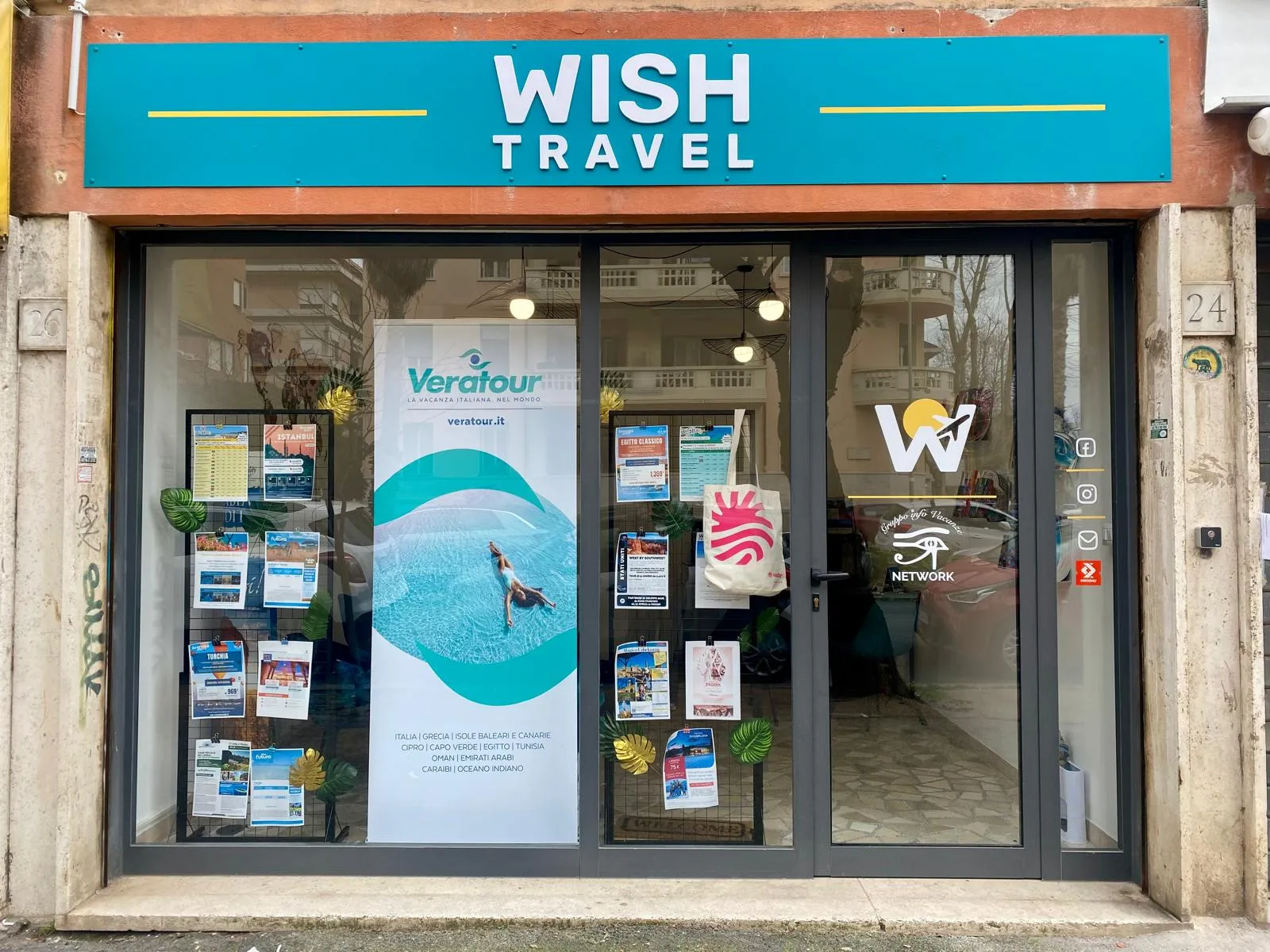 Vetrina Wish Travel con insegna turchese, banner Veratour, locandine di viaggi su griglie nere e numero civico 24